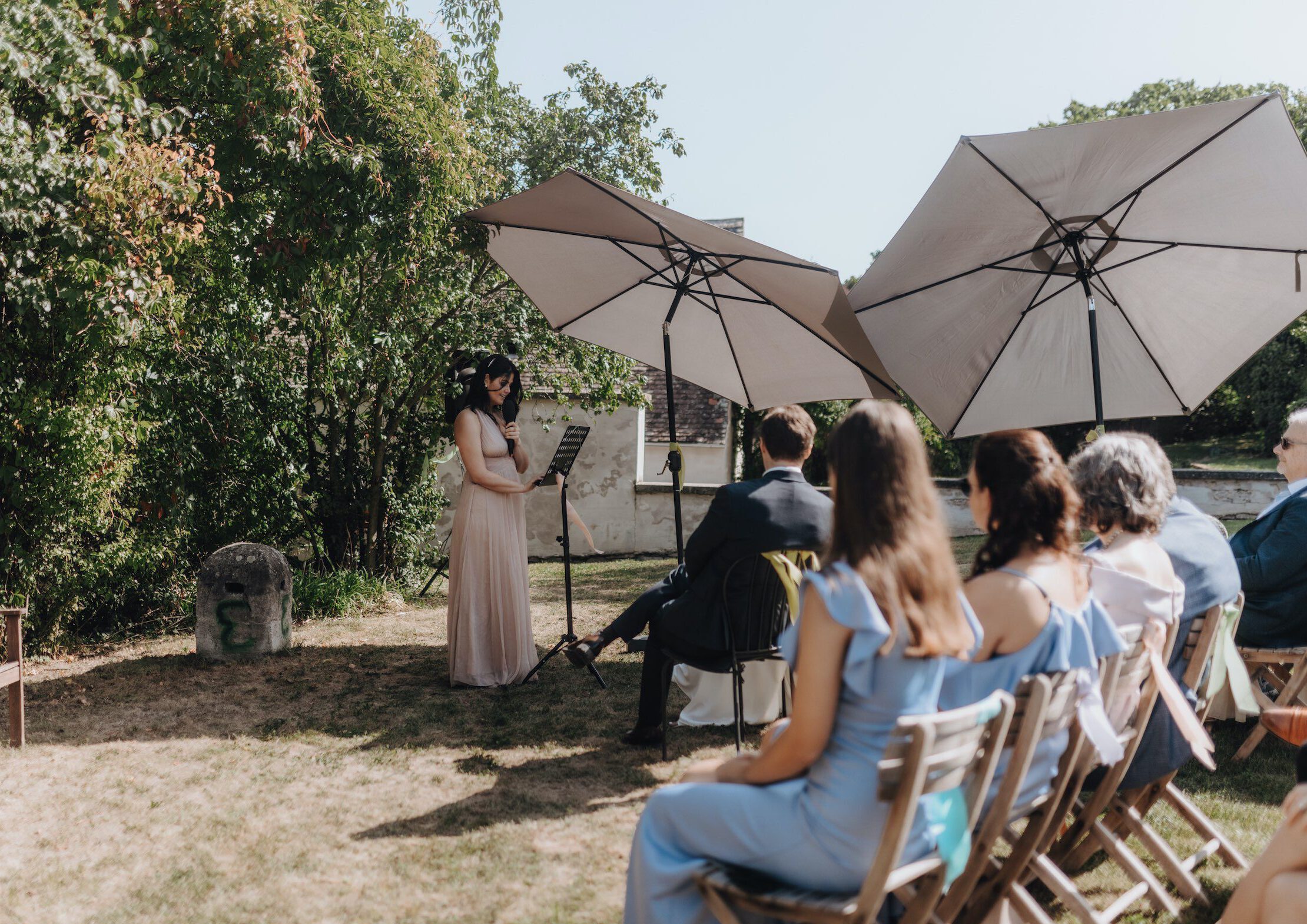 Wedding guests seated in a garden setting, listening to the ceremony under large umbrellas.